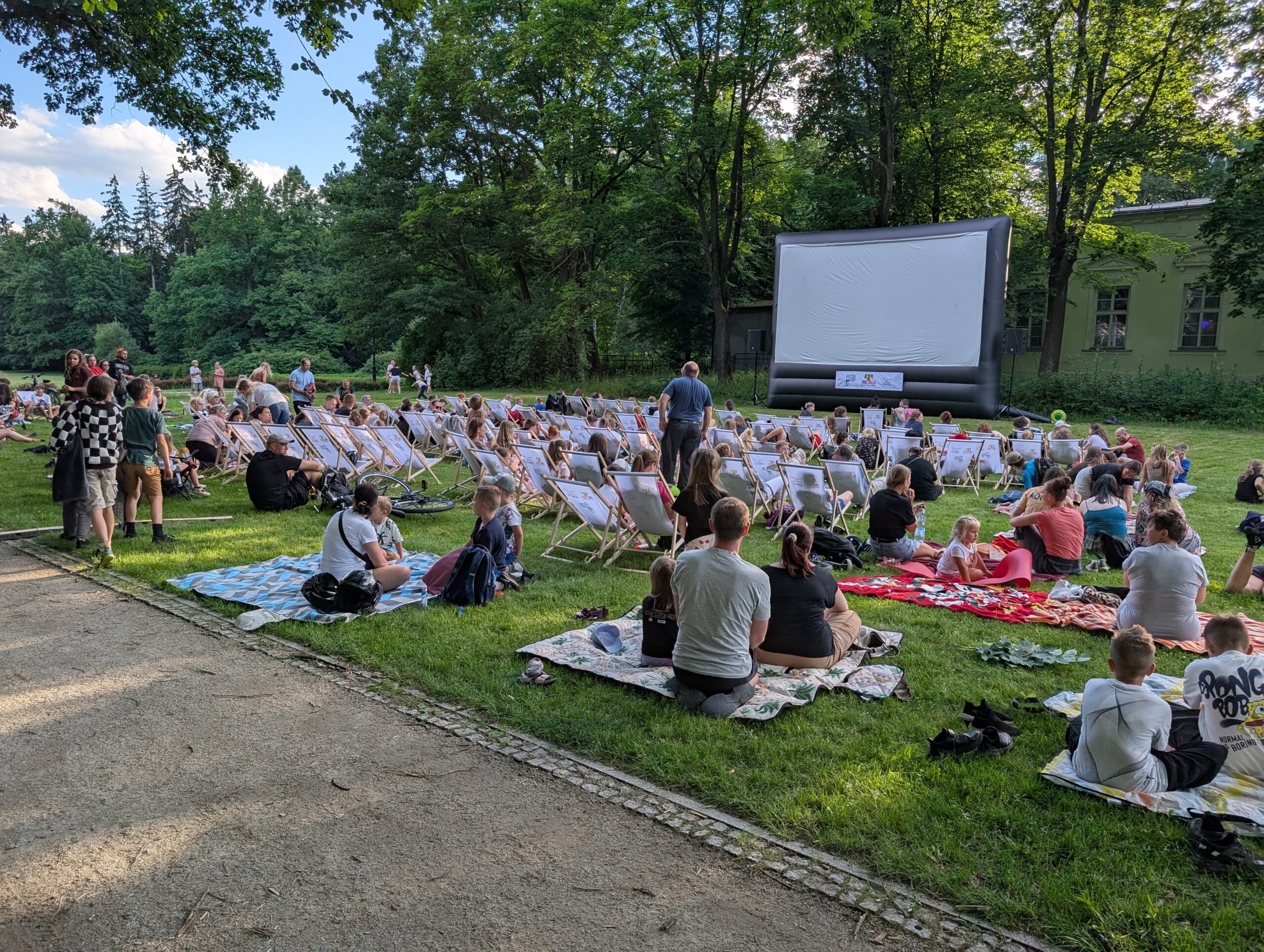 Letnie kino plenerowe w parku w Cieplicach — kilkadziesiąt osób na leżakach i kocach przed dużym nadmuchiwanym ekranem, rodziny z dziećmi, wieczorne światło